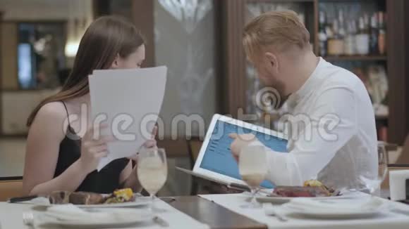 漂亮的黑发女人和金发大胡子男人坐在咖啡馆的桌子旁讨论笔记本电脑屏幕上的信息视频的预览图