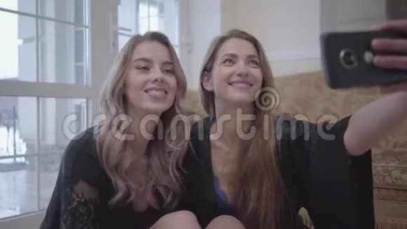 两个穿着黑色连衣裙的漂亮女人坐在家里的地板上在手机上自拍女孩子假装视频的预览图