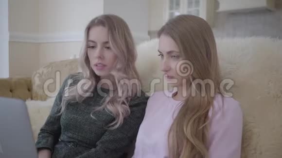 两位可爱的年轻女性正在利用坐在沙发上的笔记本电脑上网寻找优惠视频的预览图