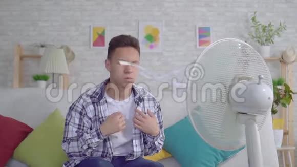 年轻的亚洲男子从坐在工作风扇前的热气中拯救了热慢的概念视频的预览图