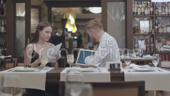 一位年轻漂亮的女士坐在餐厅的桌子上拿着文件带着一个自信的留胡子的男人向她展示视频的预览图