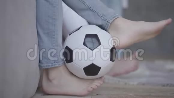 青少年男孩踢足球的脚靠近前景孩子坐在家里的沙发上模糊视频的预览图
