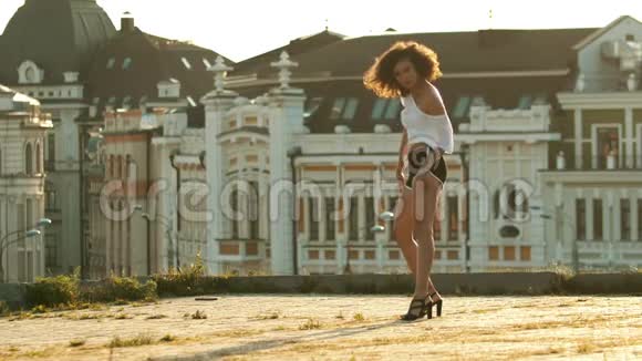 一个年轻的女人长着长腿穿着小短裤和卷发在屋顶上表演迷人的舞蹈明亮的日落视频的预览图