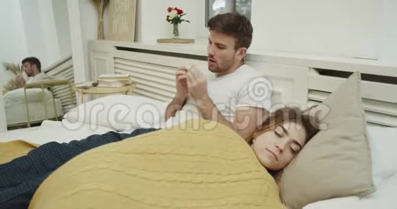 生病的男人躺在他的女人旁边躺在一张现代的床上他得了流感和流鼻涕女人在他身边睡了一觉视频的预览图
