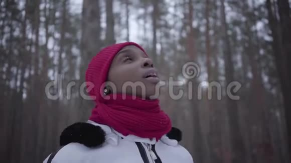 穿着温暖衣服的非裔美国女孩戴着红帽子围巾和白色夹克走在冬林里抬头望着视频的预览图