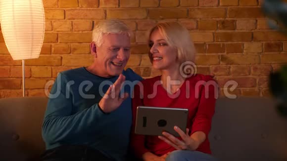 在平板电脑上播放视频的高级幸福夫妇的特写镜头他们兴高采烈地坐在室内的沙发上视频的预览图