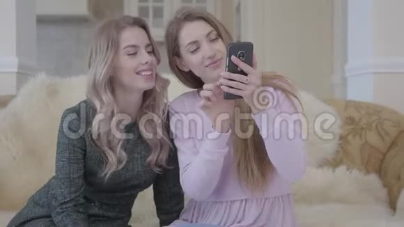两个穿着时尚服装的漂亮年轻女孩坐在客厅的沙发上看着电话寻找女孩视频的预览图