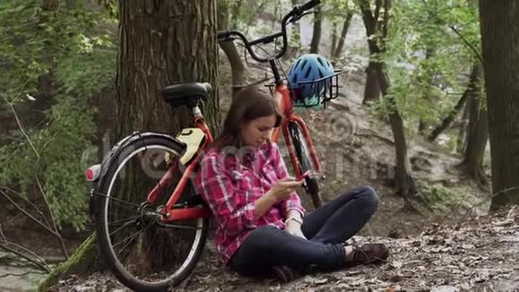生态自行车运输年轻的高加索女学生坐在公园里休息附近租着橙色自行车用手机视频的预览图