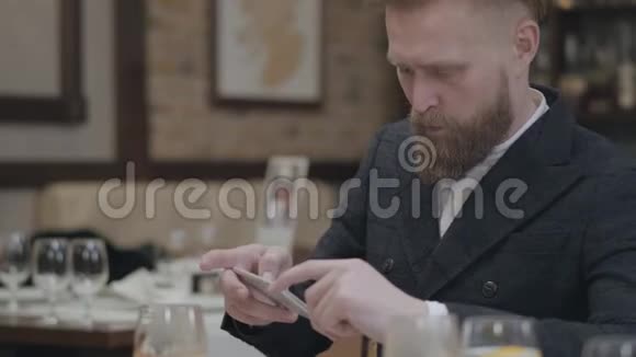 自信的金发大胡子男穿着时髦的夹克拿着手机坐在一个现代化的房间里拍照视频的预览图