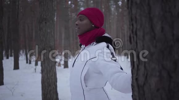 美国黑人女孩穿着温暖的衣服戴着红色的帽子围巾和白色的夹克站在冬天的森林里看着视频的预览图