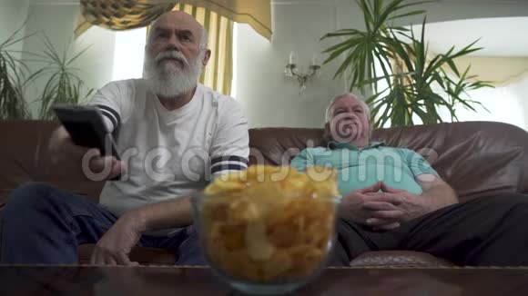 两个成熟的高级男人坐在棕色的皮革沙发上看电视老人用遥控器开关频道视频的预览图