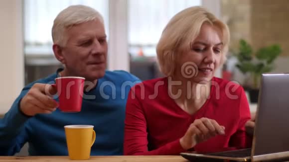在一间舒适的公寓里用笔记本电脑在桌子上和室内喝茶拍摄高级幸福夫妇的特写镜头视频的预览图