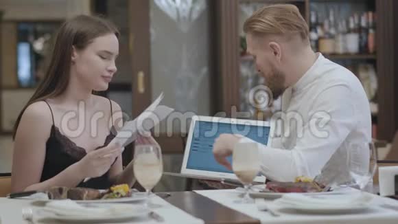 漂亮的年轻女人和金发胡子男人坐在咖啡馆的桌子旁讨论笔记本电脑屏幕上的信息视频的预览图