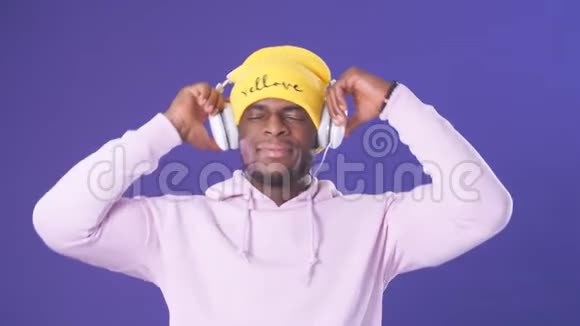 戴着帽衫和黄色帽子的深色皮肤快乐的家伙听着耳机里最喜欢的曲目视频的预览图