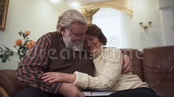 幸福的成熟夫妇坐在家里现代客厅的沙发上年长的男人和女人互相拥抱休闲视频的预览图