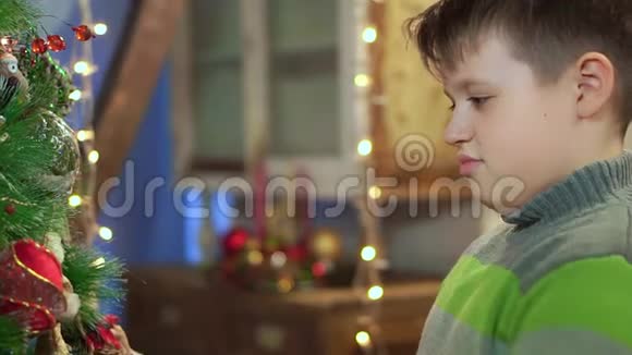 一个穿毛衣的男孩在家中的圣诞树附近检查它在圣诞灯的背景下视频的预览图