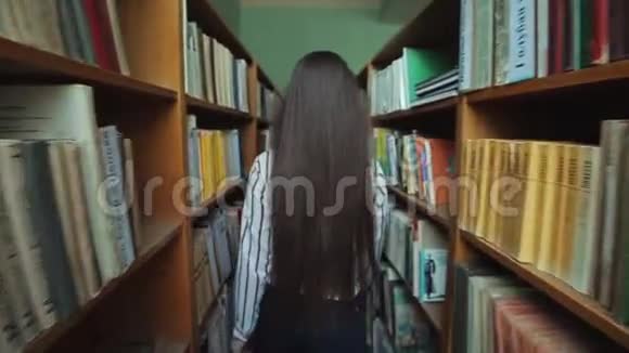 一位留着长长黑发的美丽女孩的后景在图书馆的书架之间手里拿着书慢慢视频的预览图
