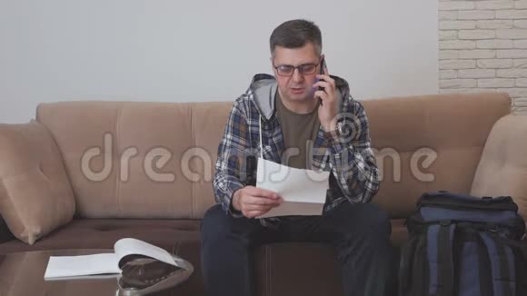 一位中年男子坐在酒店房间的沙发上拿着一份纸质文件在电话里交谈旁边是一个视频的预览图