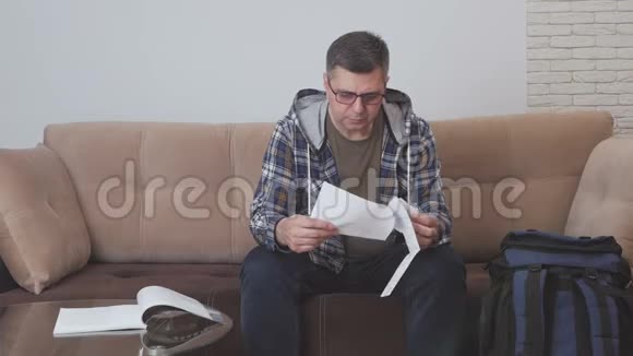一位中年男子坐在酒店房间的沙发上拿着一份纸质文件在电话里交谈旁边是一个视频的预览图