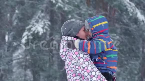 一位快乐的母亲在一个美丽的冬天雪天把她的儿子抱在树林或公园里视频的预览图