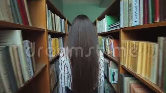 一位留着长长黑发的美丽女孩的后景在图书馆的书架之间手里拿着书慢慢视频的预览图