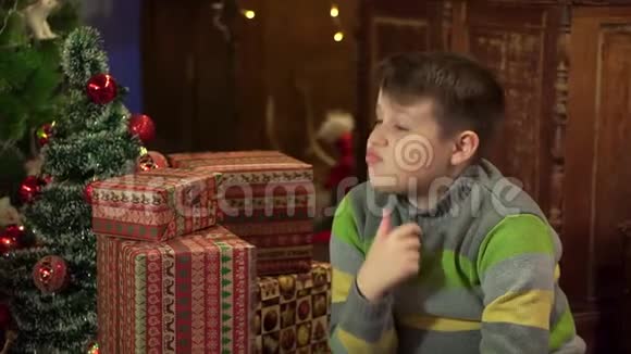 一个男孩穿着毛衣在家中圣诞树附近考虑礼物等待在圣诞灯的背景下视频的预览图