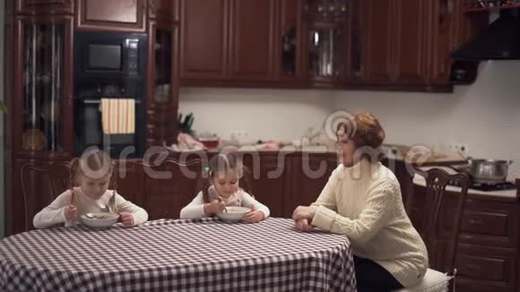 奶奶和两个可爱的孙女坐在桌子旁的大猫子里吃汤幸福的家庭在家吃午饭视频的预览图