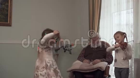 大胡子老人坐在大窗户附近的摇椅上看着他跳舞的孙女她姐姐站在视频的预览图