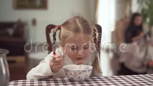 可爱的金发女孩扎着辫子在模糊的背景下坐在现代厨房的桌子旁吃着罗什小女孩视频的预览图