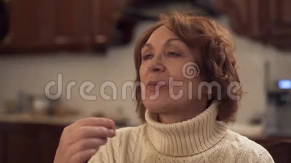 穿暖和毛衣的老妇人在现代厨房里坐在家里吃糖果然后露出大手指成熟的女士享受视频的预览图
