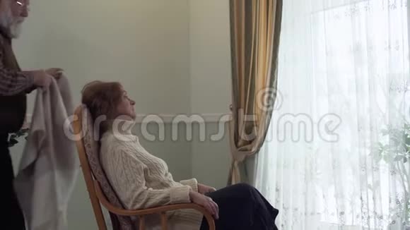 成熟的女人坐在现代房子大窗户附近的摇椅里她丈夫来给妻子盖上毯子视频的预览图