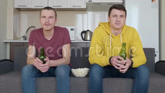 两个男人坐在沙发上看电视上的足球赛喝啤酒吃爆米花视频的预览图