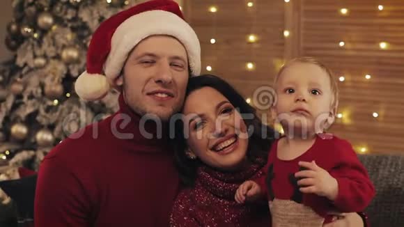 圣诞节那天幸福的年轻家庭坐在沙发上在家里的休息室里微笑着看着视频的预览图
