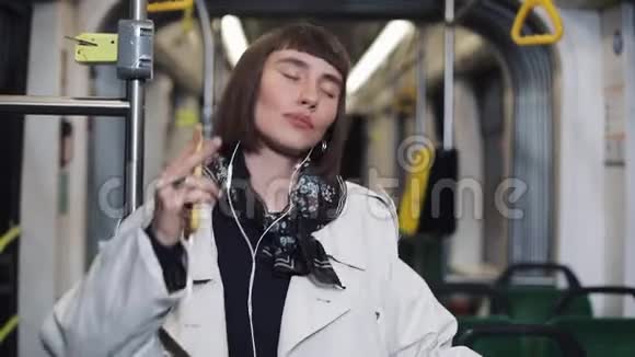 年轻快乐的女人穿着外套戴着耳机听音乐和在公共交通中有趣的舞蹈的肖像视频的预览图