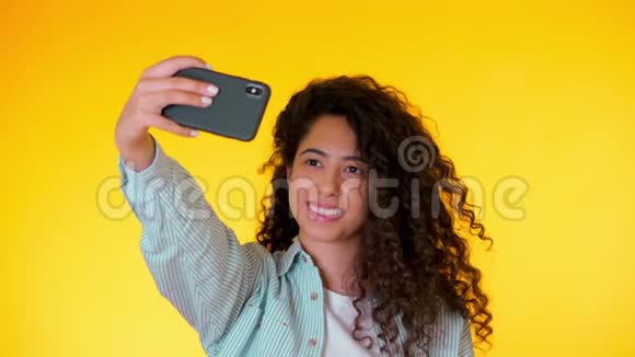 美丽的女人卷发拿着智能手机在黄色的工作室里拍摄自己使用技术的女性视频的预览图