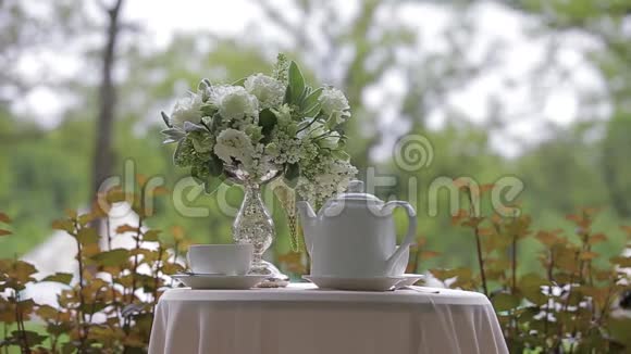 热茶白色的杯子和白色的茶壶提供在户外白色的桌子装饰着一束美丽的花视频的预览图