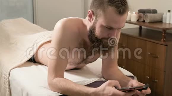 留着辫子的胡子男子躺在医疗沙发上在办公室的智能手机上发送按摩短信视频的预览图