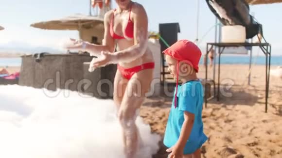 在肥皂海滩迪斯科舞厅里快乐的年轻女子用肥皂泡沫与她蹒跚学步的儿子玩耍视频的预览图