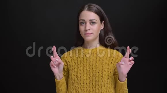穿黄色毛衣的长发黑发女孩的肖像示意交叉手指标志以显示黑色背景上的希望视频的预览图