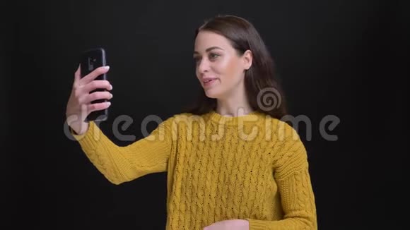 穿着黄色毛衣的长发黑发女孩用黑色智能手机在电视上愉快地交谈的肖像视频的预览图