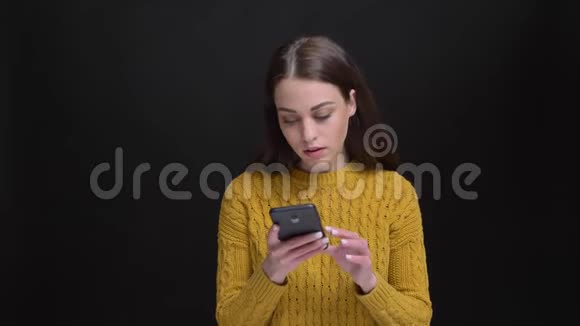 一幅长头发的黑发女孩穿着黄色毛衣专注地在黑色背景下与智能手机合作的肖像视频的预览图