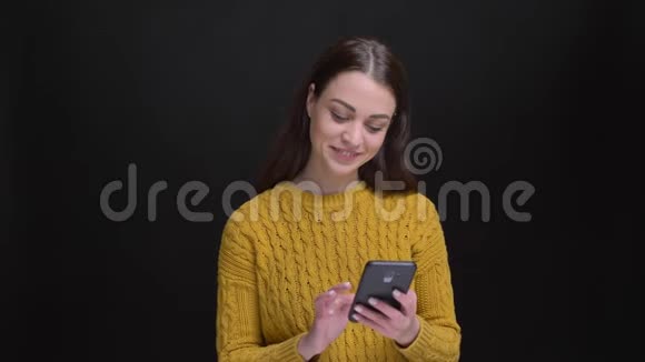 一幅长发黑发女孩穿着黄色毛衣在黑色背景下愉快地与智能手机合作的肖像视频的预览图