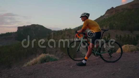 年轻的运动员坐在山顶上的自行车上戴着黄色的t恤头盔和运动器材看着视频的预览图