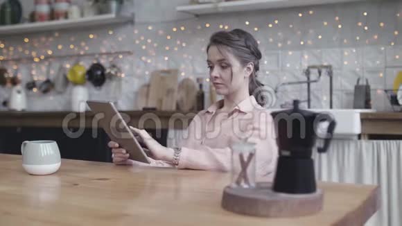 严肃美丽的女士坐在厨房里的木米黄色桌子旁用她的iPad浏览一些东西股票视频的预览图