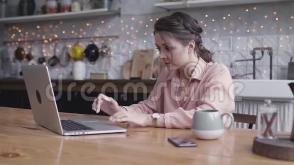 可爱的黑发女人坐在厨房的一张桌子旁用手提电脑在网上工作沉思视频的预览图