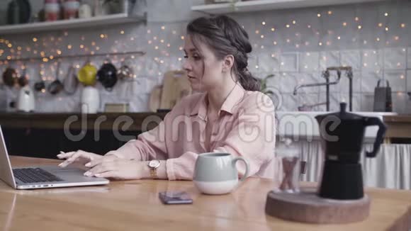 一个年轻的女人坐在厨房的桌子上用笔记本电脑库存录像在家工作的漂亮黑发女人视频的预览图