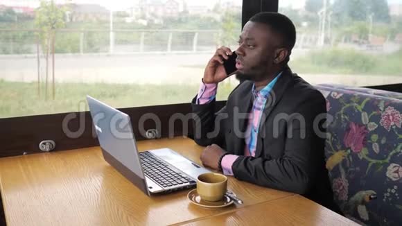 黑人商人在笔记本电脑上工作打电话给智能手机坐在咖啡屋里视频的预览图
