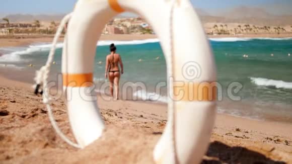 4K镜头性感年轻女子穿迷你比基尼在沙滩上散步在风日相机寻找拯救生命视频的预览图