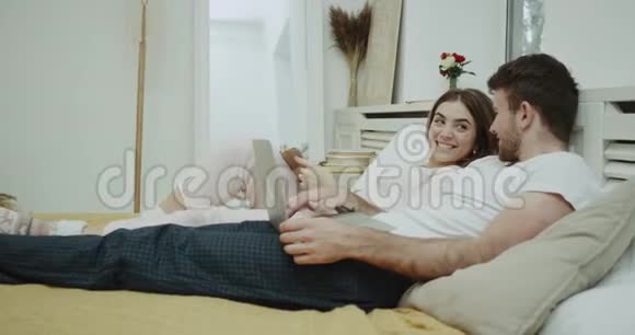 一对夫妇在床上的早晨例行公事女人读一本书她的伴侣在笔记本电脑上看拍摄红色史诗视频的预览图