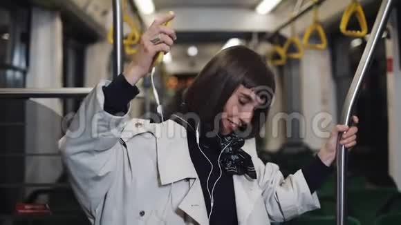 年轻女子穿着外套戴着耳机听音乐和在公共交通工具上滑稽跳舞的肖像他是视频的预览图
